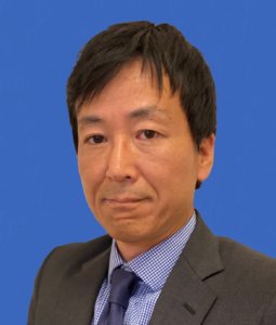 professional headshot of japanese male wearing a black suit jacket and blue tie against a blue background